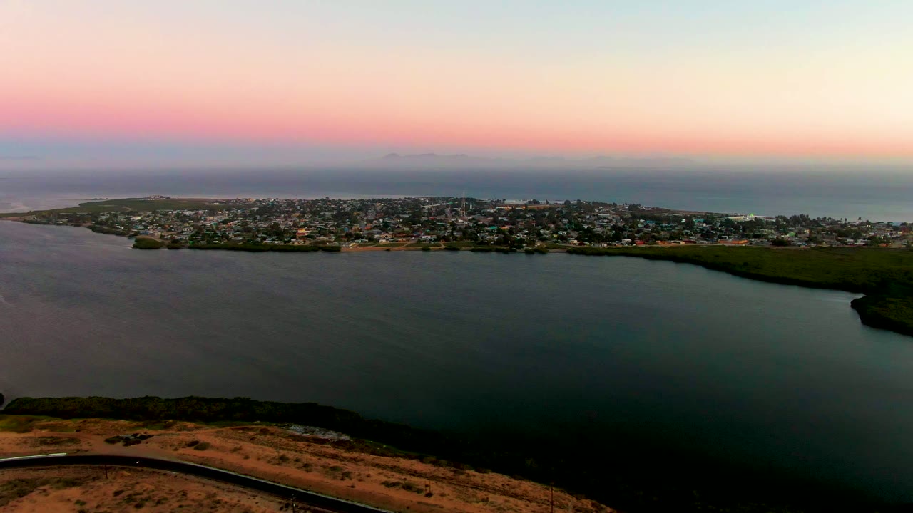 Aerial dusk view over San Carlos, Baja California Sur, with a coastal road at the bottom, calm water in the middle, a town on the peninsula, ocean beyond, and no visible people.