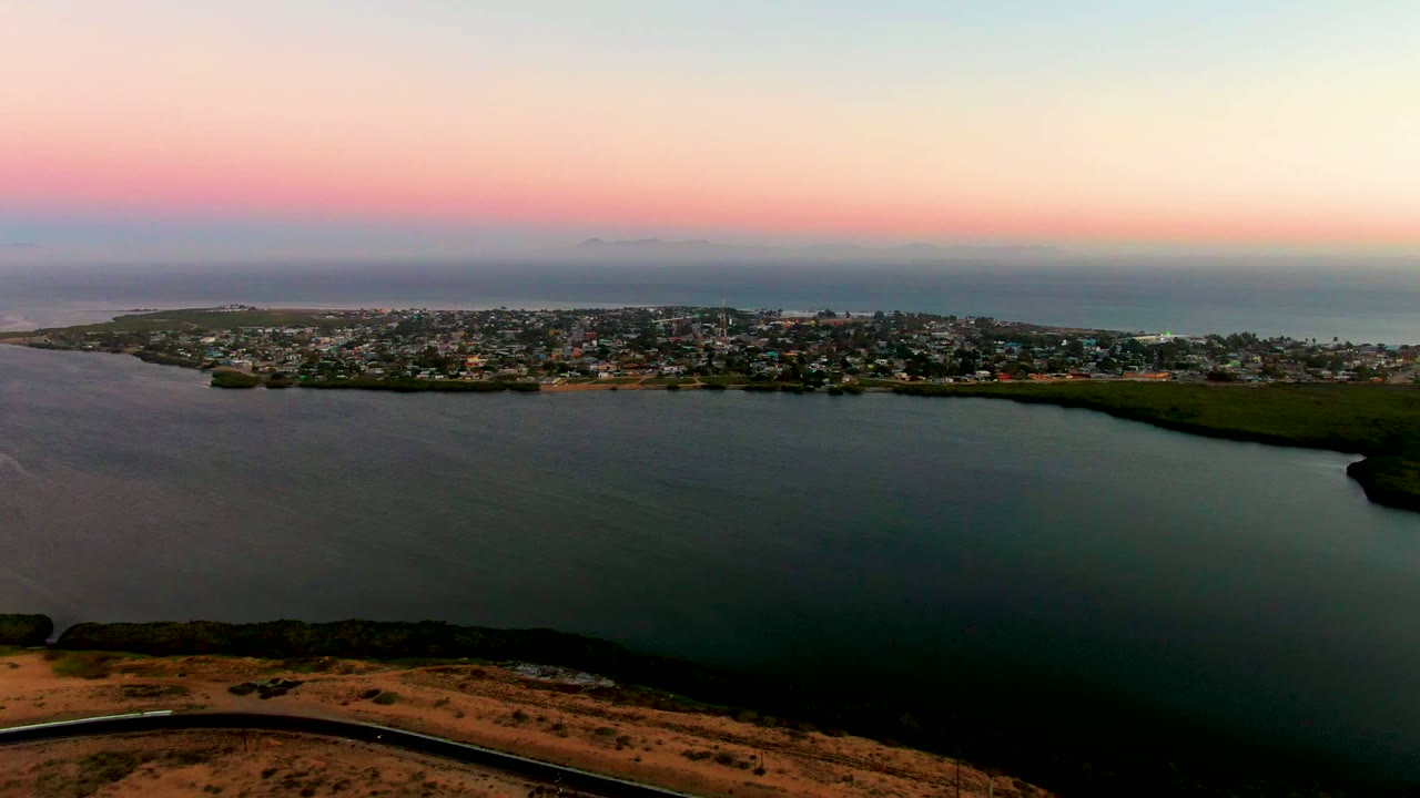 Wide aerial frame over San Carlos, Baja California Sur, with a small vehicle on the low foreground road, dark calm water, low-rise town buildings, ocean horizon, and no visible people.
