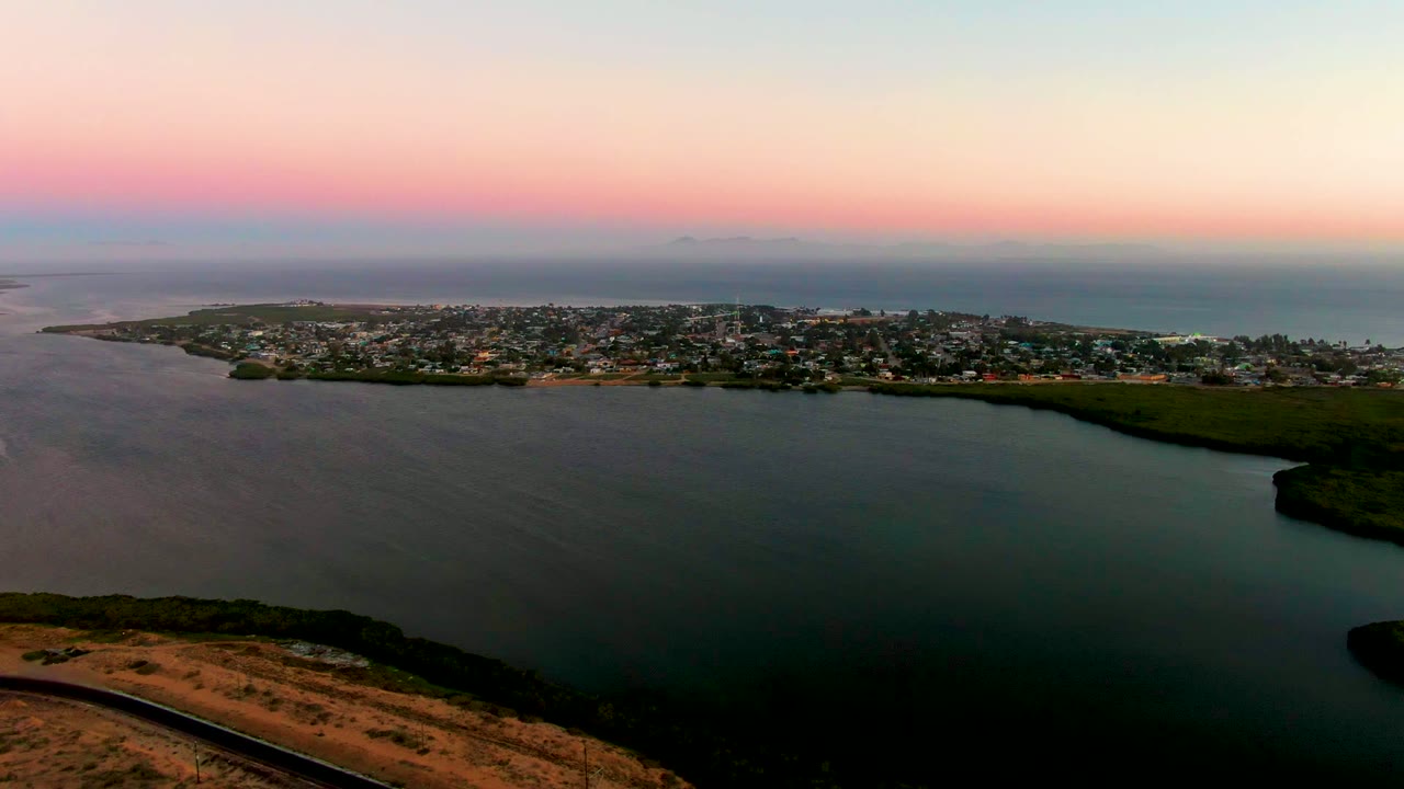 Aerial dusk view over San Carlos, Baja California Sur, with a dry hillside road in the foreground, calm bay water, a coastal town beyond, ocean on the horizon, and no visible people.