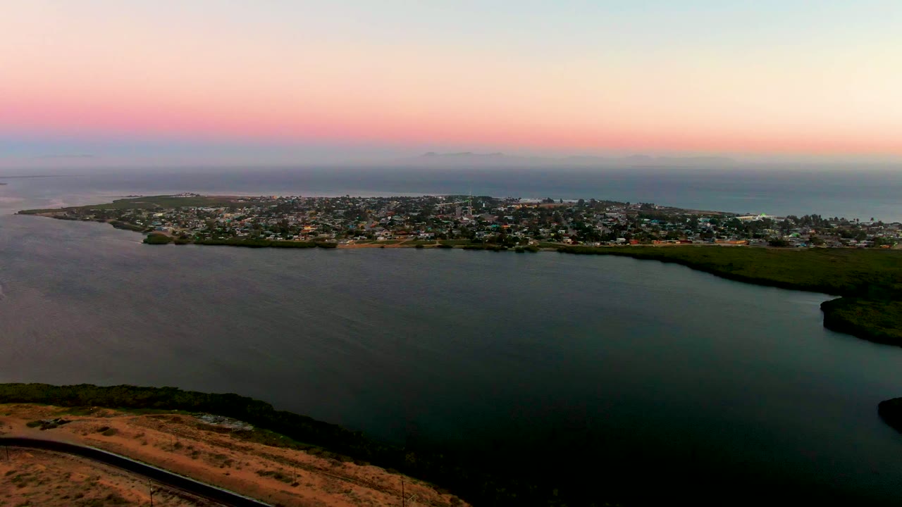 Wide aerial view of bay water, coastal homes, hillside road, and sunset sky.