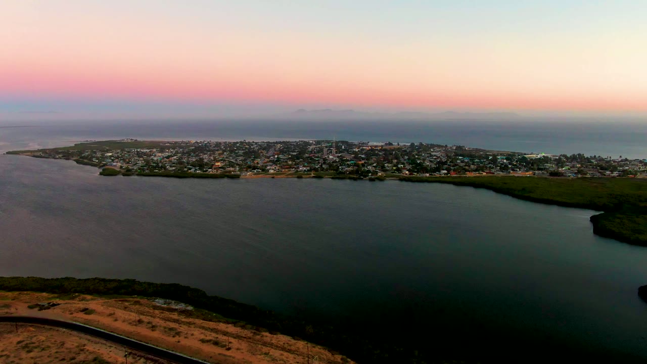 Aerial view across the bay toward San Carlos, Baja California Sur, with the town stretched along the shoreline, dark water filling the foreground, distant ocean behind it, and no visible people.