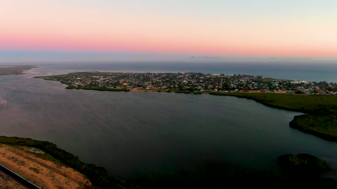 Aerial dusk view in San Carlos, Baja California Sur, looking across a dark bay toward a low coastal town and ocean horizon under pink sky, with no visible people.