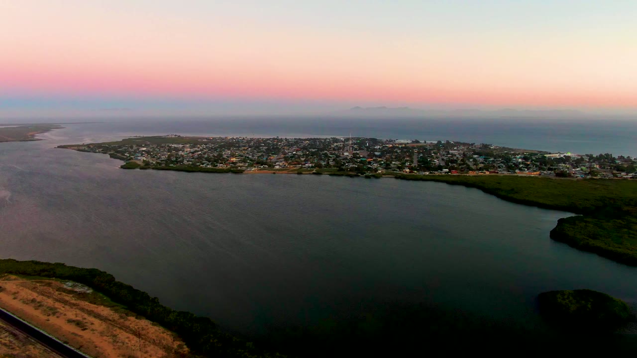 Wide aerial of bay water, shoreline greenery, and town beneath a pink horizon.