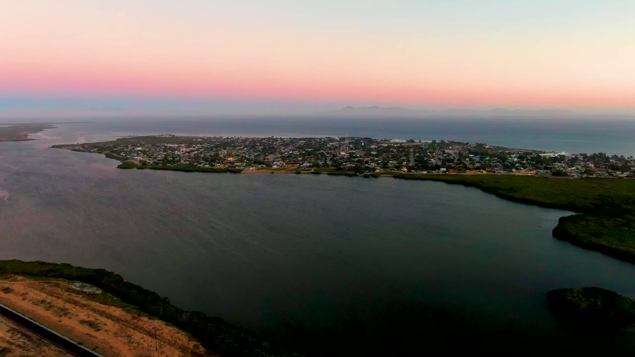 San Carlos, Baja California Sur appears across a sheltered bay, with dark water in the foreground, a green shore on the right, ocean beyond town, and no visible people.