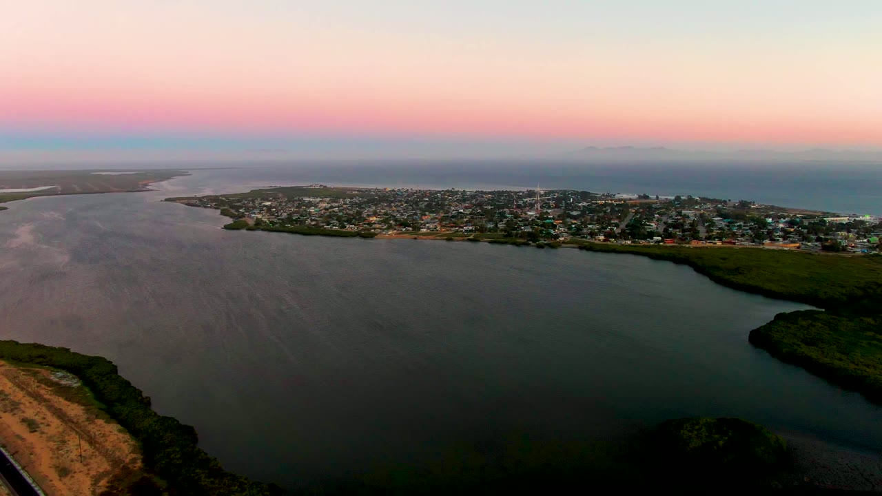 At 0:00, San Carlos, Baja California Sur appears from above with dark bay water in front, low shoreline housing across the middle, open ocean beyond, and no visible people.