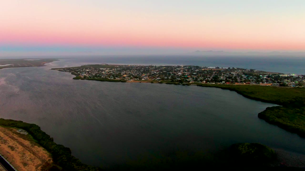 Wide drone view over calm bay water toward a coastal town and ocean horizon.