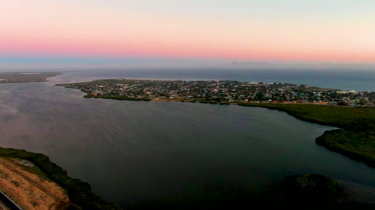 At 0:02, San Carlos remains centered along the shoreline while darker bay water fills the foreground, open ocean sits behind the buildings, and no visible people appear.