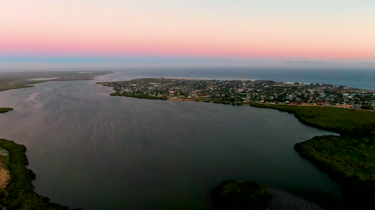 Opening aerial frame over San Carlos, Baja California Sur, with broad lagoon water in front, a coastal town across the center, ocean on the horizon, pink dusk light, and no visible people.