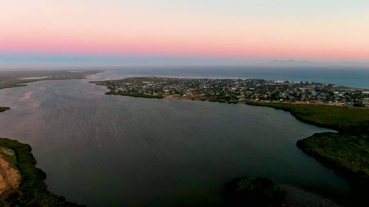 Aerial frame from San Carlos with lagoon water stretching across the foreground, town buildings forming a thin band, a green point of land at right, ocean beyond, and no visible people.