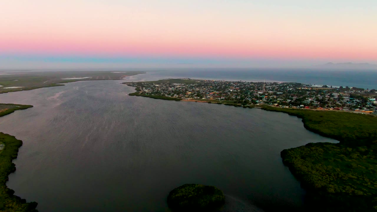 Wide aerial frame over San Carlos, Baja California Sur, showing dark lagoon water, green shoreline vegetation, a coastal town across the water, distant sea, pink dusk sky, and no visible people.