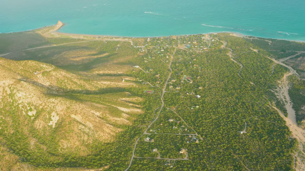 Early frame of Cabo Pulmo coastal hills in Baja California Sur, with dry tan slopes, green vegetation, pale roads, scattered buildings, turquoise sea beyond, and no visible people.