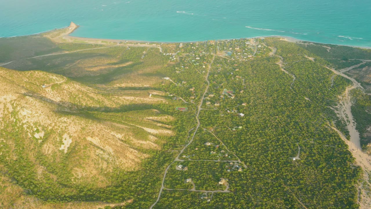 Aerial coastal hillside with shadowed terrain, roads, buildings, and ocean horizon.