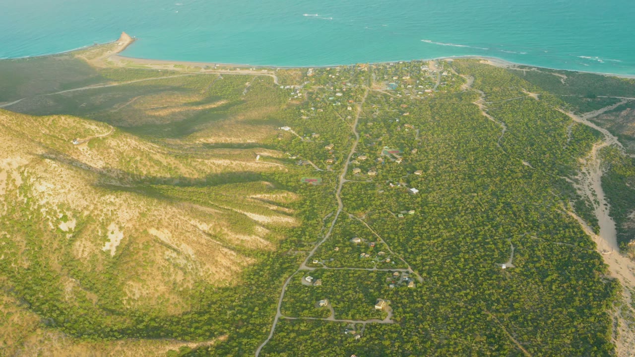 Mid-clip aerial view in Cabo Pulmo with green vegetation dominating the lower right, dry ridges to the left, road lines through the hills, sea at the top, and no visible people.