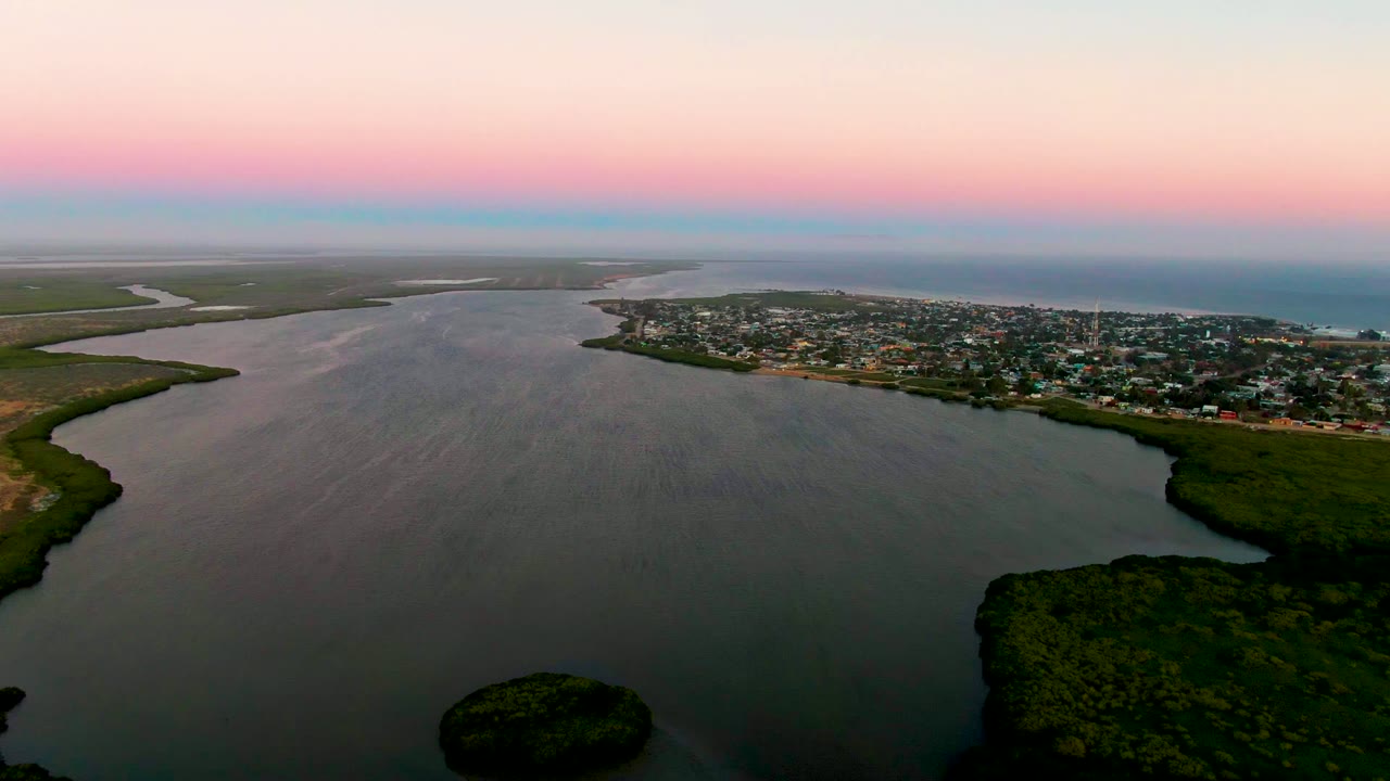 Wide drone view of calm water, marshy shoreline, town, and pastel evening sky.