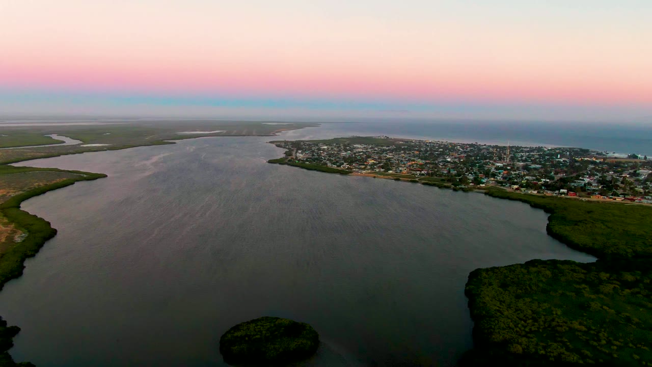 Aerial landscape over San Carlos, Baja California Sur, with darker foreground water, a green shoreline point at right, distant houses across the water, a pink horizon band, and no visible people.