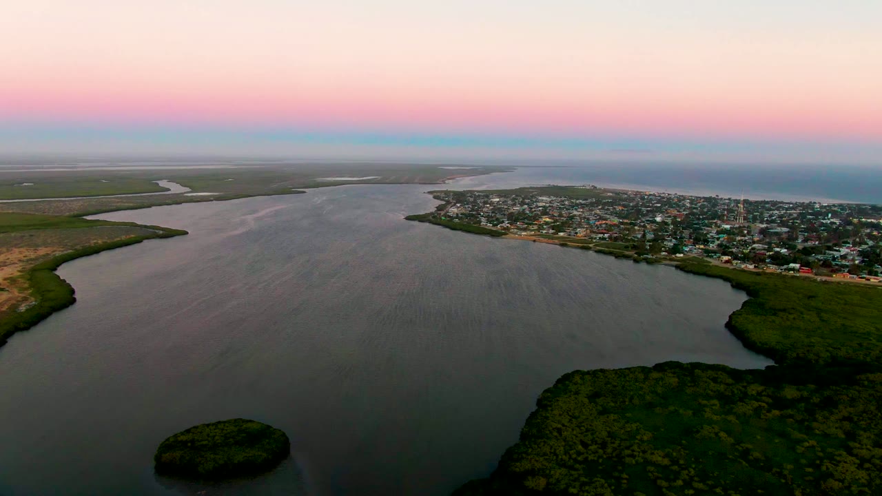 Aerial view over San Carlos, Baja California Sur at dusk, with a small island in the lower water, green estuary vegetation, town buildings on the right shore, distant coastal water, and no visible people.