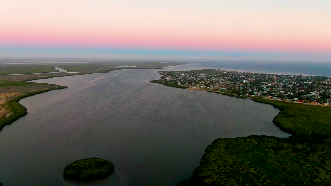 Wide aerial estuary scene with rippled water, marshy shorelines, and town buildings at dusk.