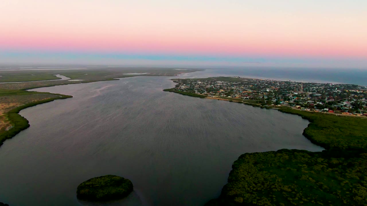 Aerial frame over San Carlos with the dark green island near the lower center, broad rippled estuary water, a populated shoreline to the right, open water beyond, and no visible people.