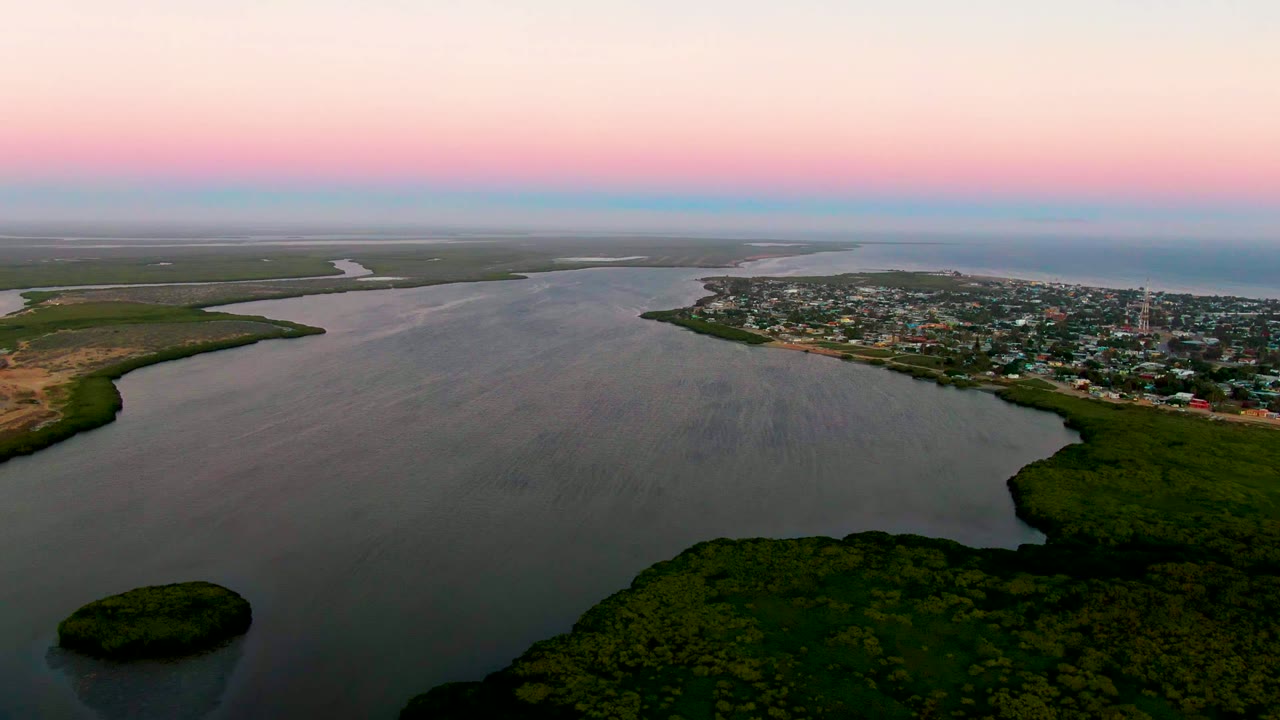 Aerial dusk view over San Carlos, Baja California Sur, with a broad river curving past green vegetation and a coastal town under a pink and blue sky, no visible people.