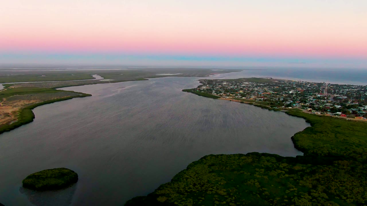 Wide aerial view of calm water, green shoreline, and town under pink dusk light.