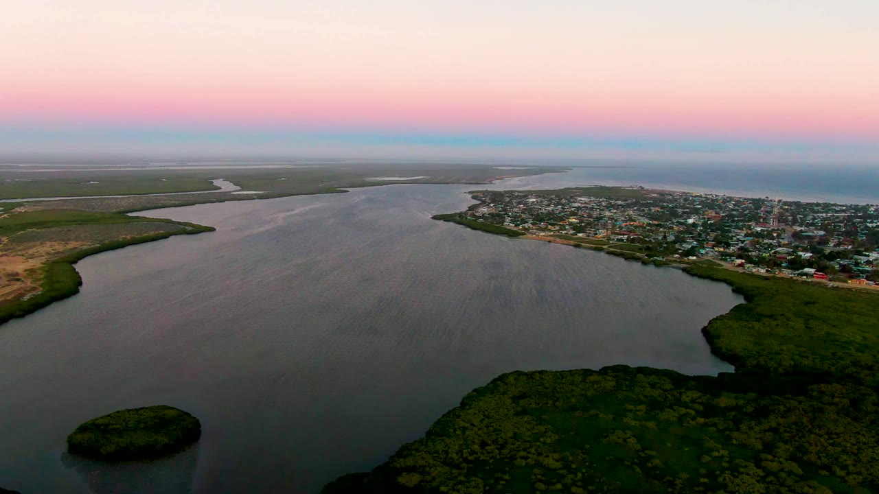 A high aerial frame over San Carlos, Baja California Sur shows the river bend, a small vegetated island near the lower left, the town along the far bank, and a pastel dusk horizon, no visible people.
