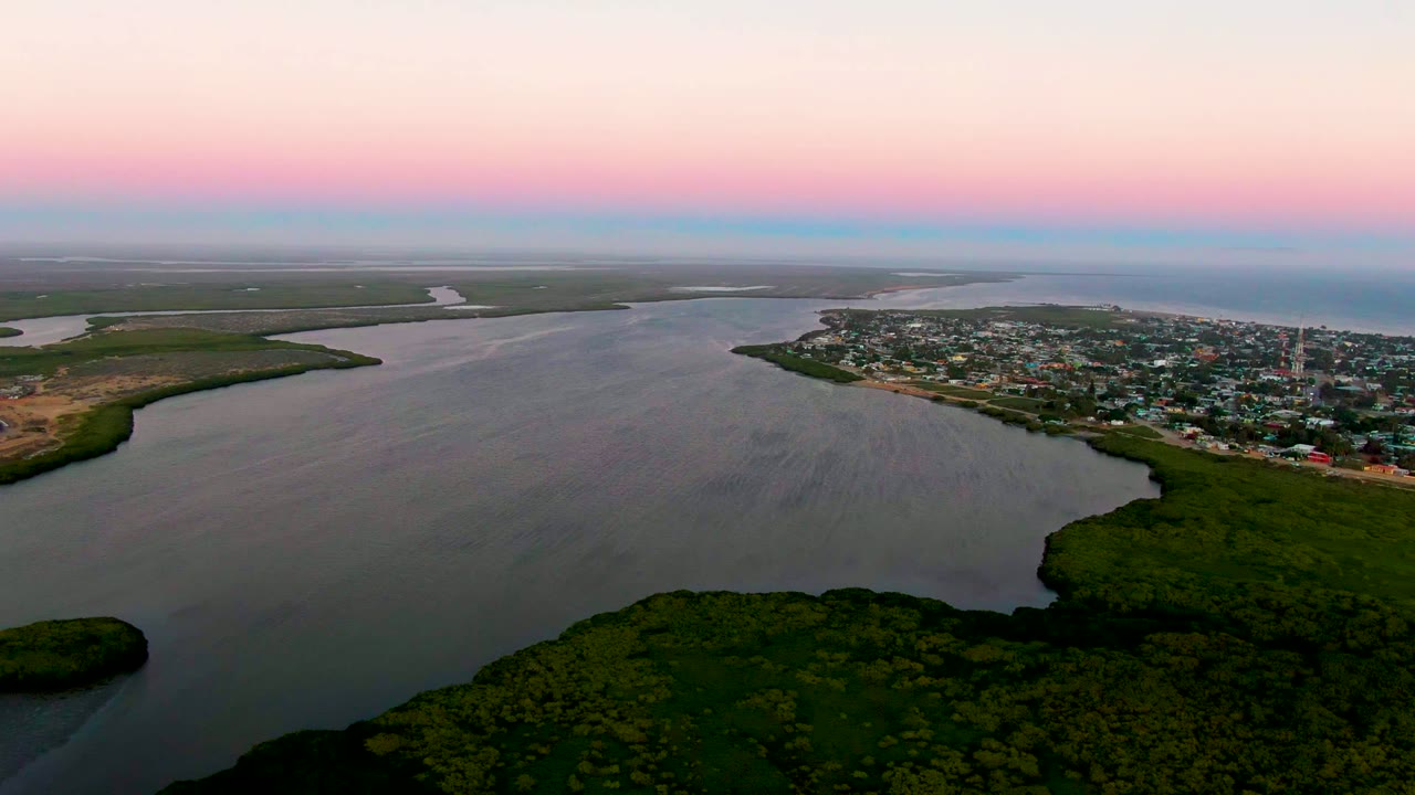 Aerial dusk view over San Carlos, Baja California Sur, with dark green wetlands in the foreground, a curving river, a low town on the right bank, and no visible people.
