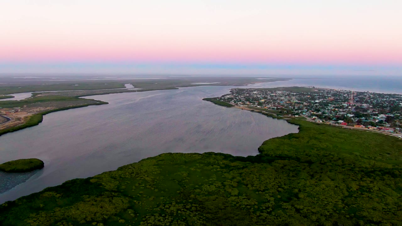 Aerial twilight view over San Carlos, Baja California Sur, with dark green wetlands in the foreground, rippling estuary water, a small town across the shore, and no visible people.