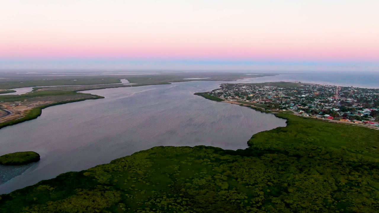 Wide aerial frame of estuary water, green shoreline vegetation, and distant town buildings beneath a pink sky.