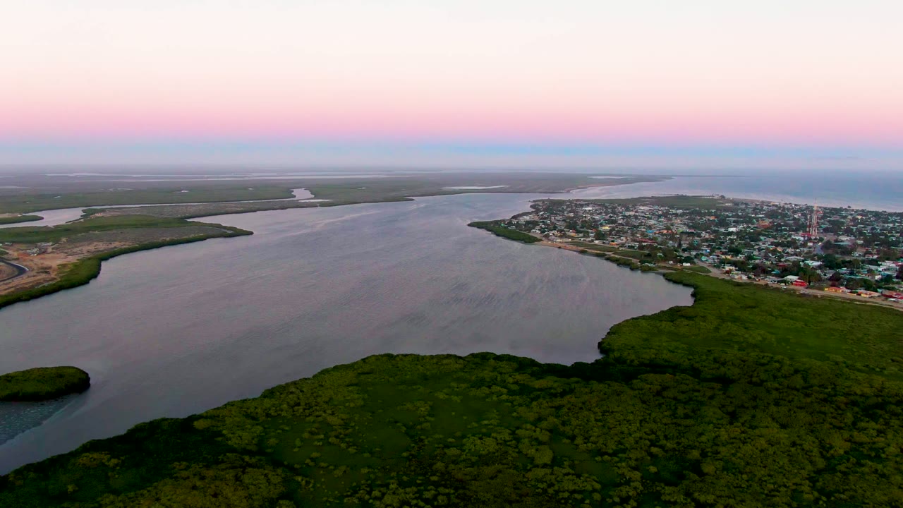Aerial dusk frame from San Carlos, Baja California Sur, with the estuary curving around green wetland banks, clustered town structures beyond the water, and no visible people.