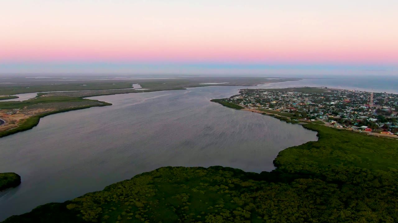Opening aerial frame of San Carlos, Baja California Sur, with dark green forest in the foreground, broad estuary water, a shoreline town, pink dusk sky, and no visible people.