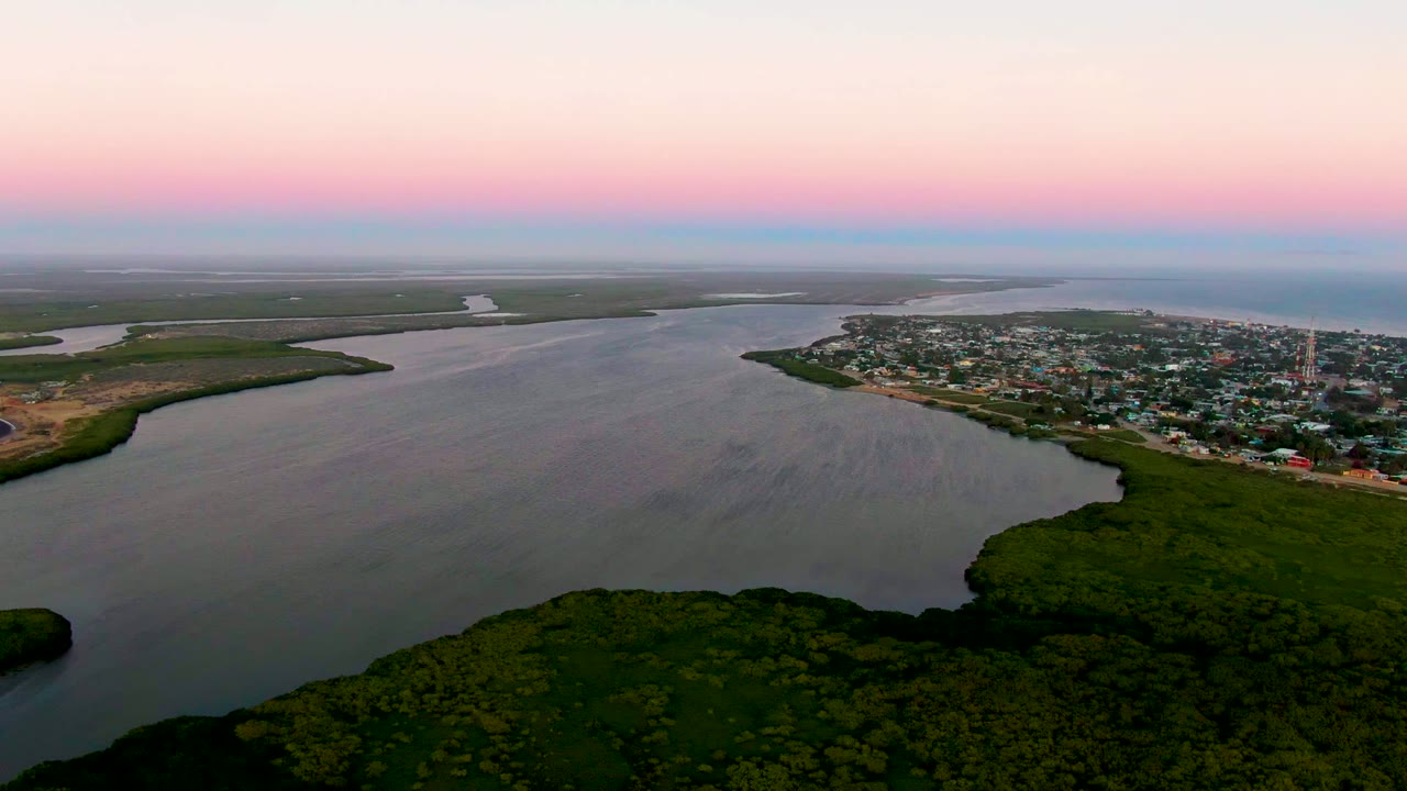 Aerial frame of San Carlos with the small town stretching along the right shore, dark foreground vegetation, calm estuary water, flat wetlands beyond, and no visible people.