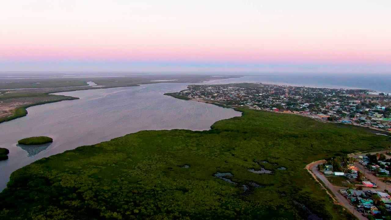 Wide drone view of San Carlos at dusk with dark green foreground forest, broad river water, coastal buildings on the right, distant open water, and no visible people.
