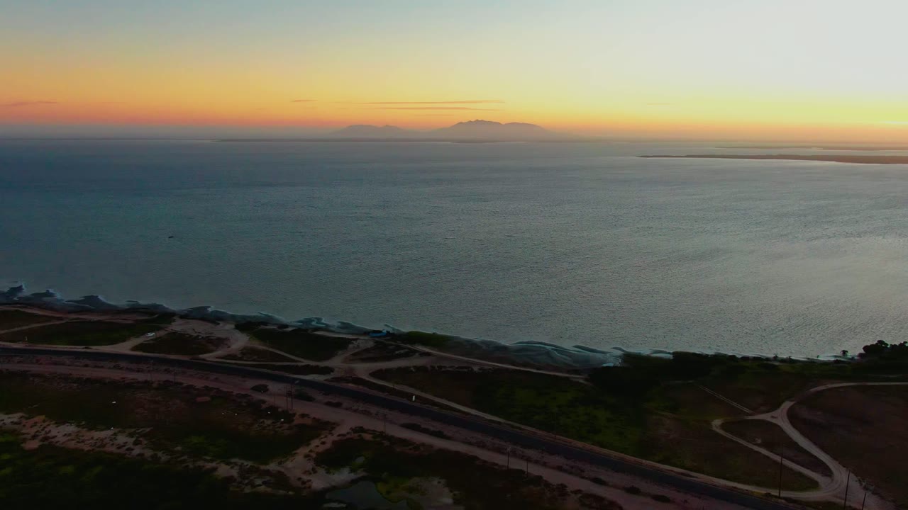 Aerial view in San Carlos, Baja California Sur showing dark coastal roads below, open ocean, distant mountains on the horizon, orange twilight sky, and no visible people.