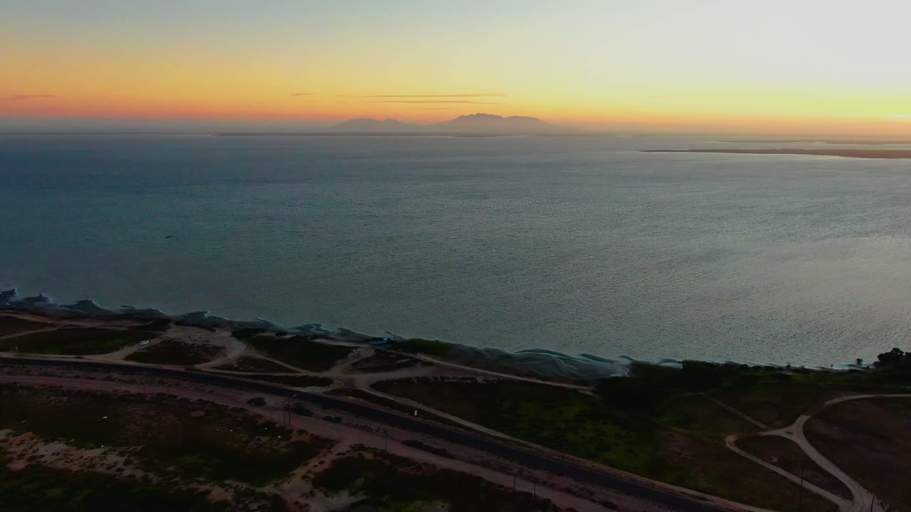 Wide aerial coastline with winding roads below and orange sky above the ocean.