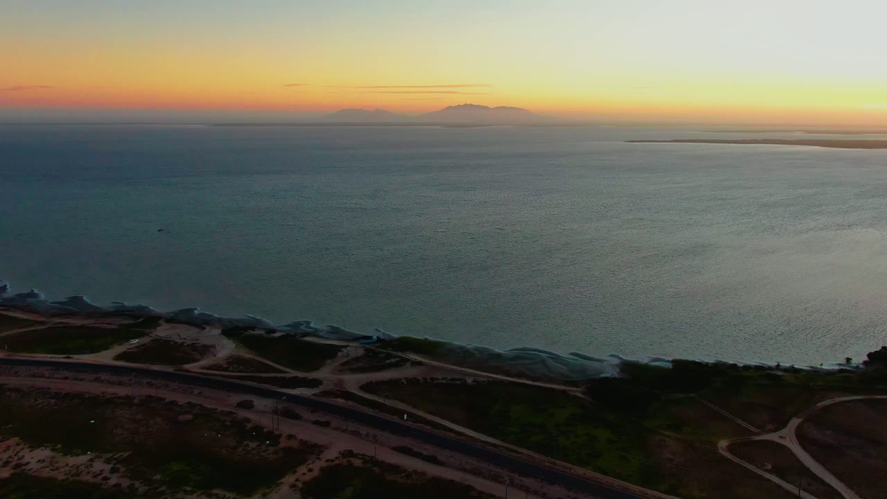 San Carlos coastal aerial showing curving road lines and dark shore in the foreground, calm ocean beyond, distant mountains left of center, and no visible people.