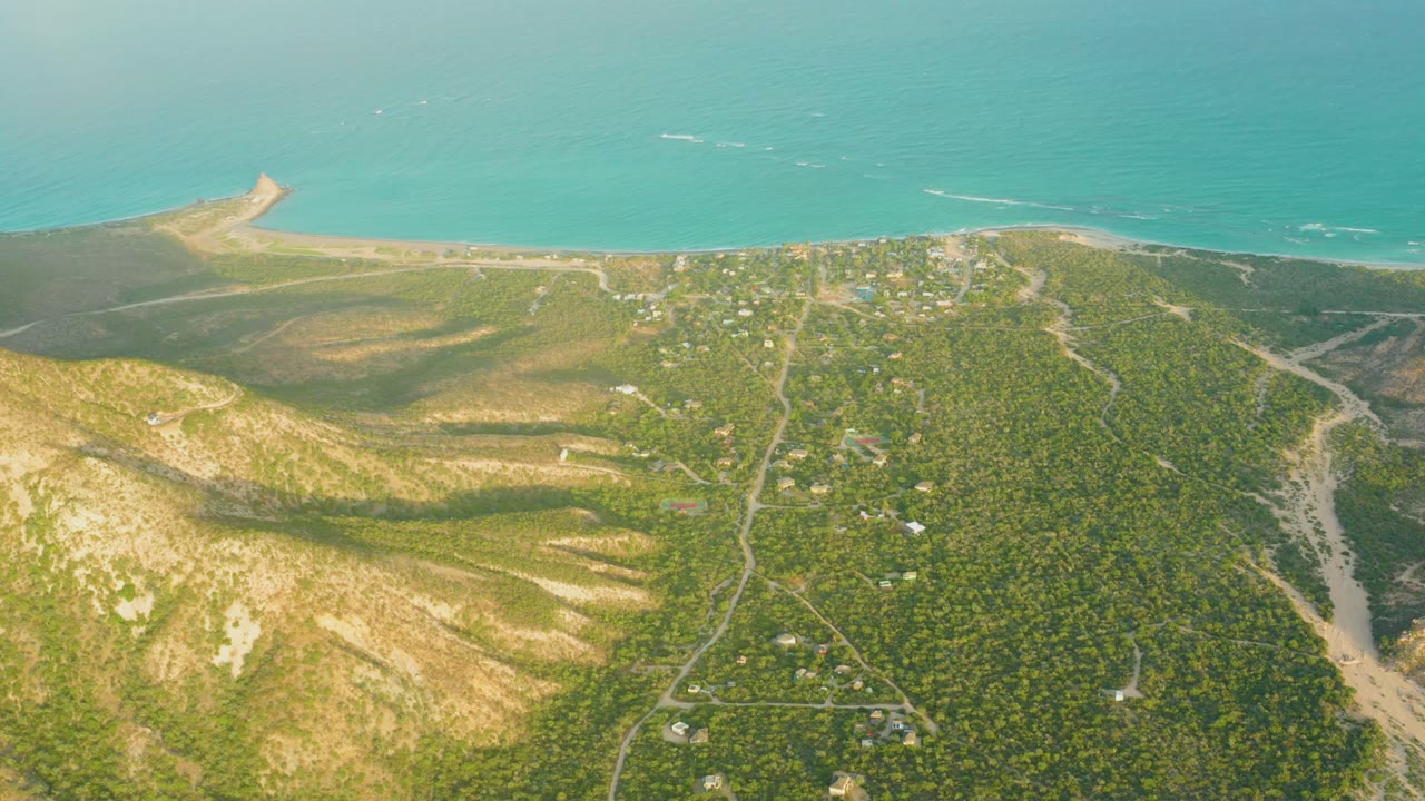 High aerial frame over Cabo Pulmo, Baja California Sur, showing green hills, sandy ridges, scattered roads, a beach strip, turquoise ocean, and no visible people.