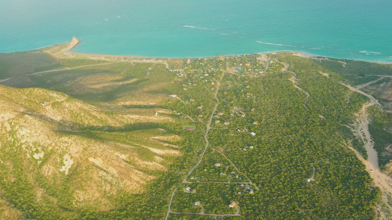 Drone frame over Cabo Pulmo showing a broader spread of green hillside roads and small structures below a pale beach and turquoise ocean, with no visible people.