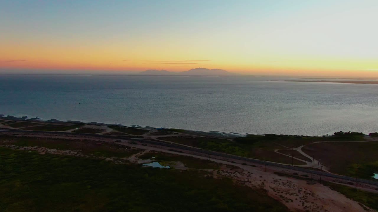 At 0:00 in San Carlos, Baja California Sur, a high aerial view shows a winding coastal road below dark dunes, broad ocean water, distant mountains on the horizon, and no visible people.
