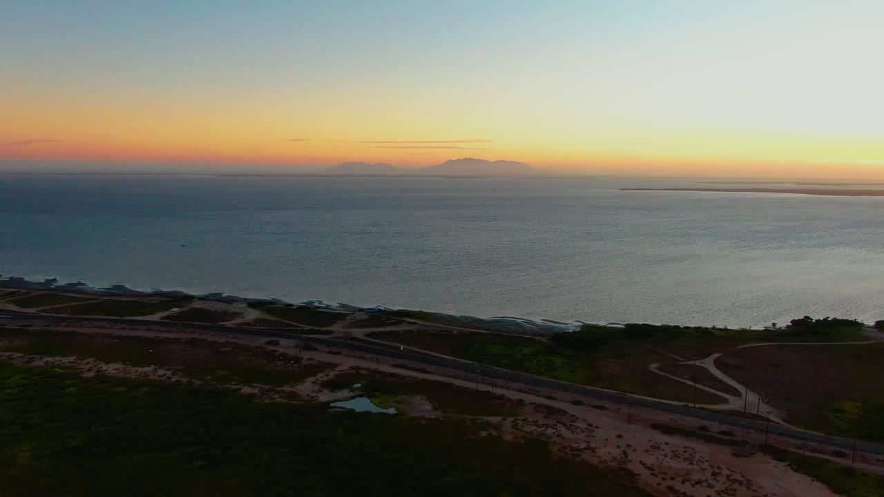 Coastal highway and dune paths below a sunset ocean horizon.