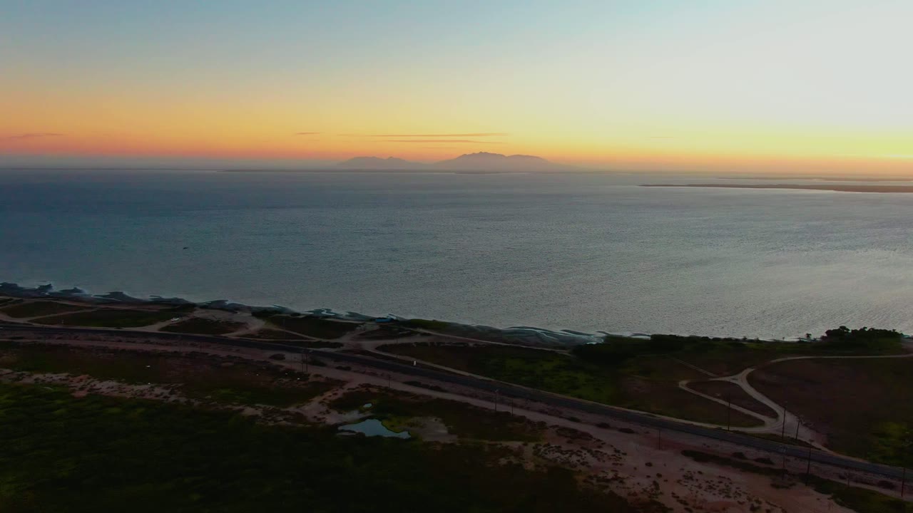 At 0:02 in San Carlos, Baja California Sur, road curves and sandy dunes remain visible in the foreground as the ocean stretches to distant mountain silhouettes beneath a warm sunset band, with no visible people.