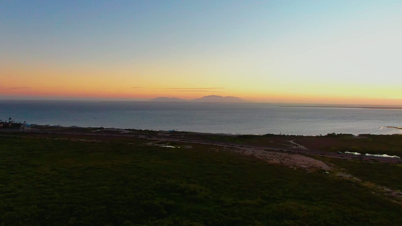 Wide aerial view in San Carlos, Baja California Sur showing dark green coastal land, pale roads, ocean horizon, distant island silhouettes, warm sunset sky, and no visible people.