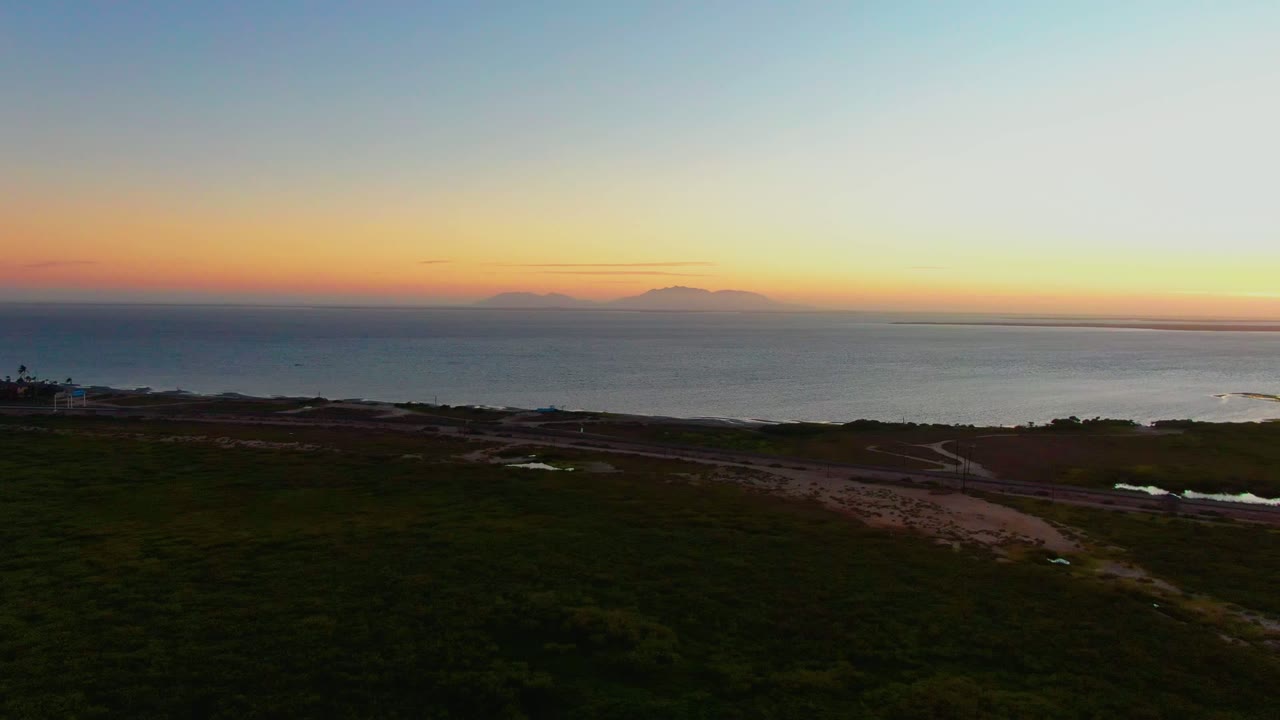 Aerial sunset frame in San Carlos, Baja California Sur with foreground roads crossing dark coastal vegetation, ocean water in the midground, distant islands on the horizon, and no visible people.