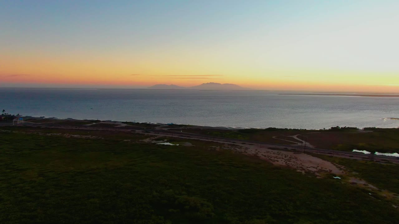 San Carlos coastal sunset view with ocean stretching to distant islands, warm light along the horizon, dark undeveloped land and roads below, a small structure near the left shoreline, and no visible people.