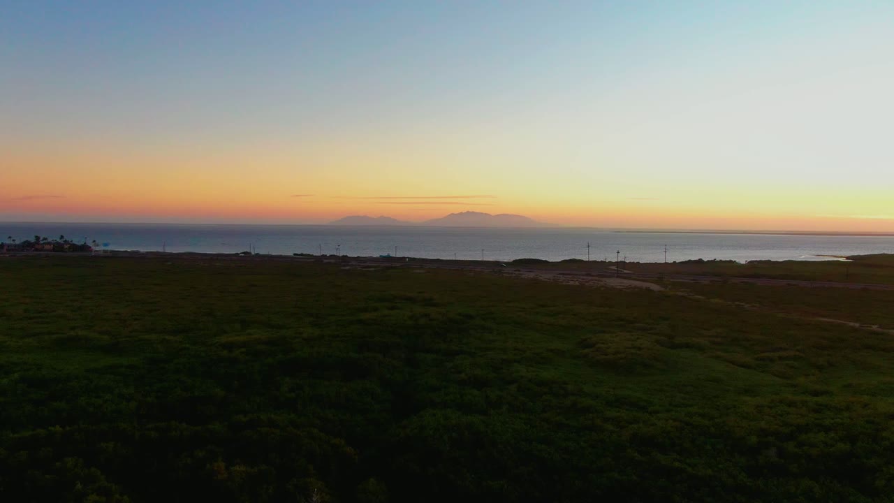 Wide elevated view of dark coastal grassland in San Carlos, Baja California Sur, with the ocean horizon and orange-blue sunset sky beyond, no visible people.