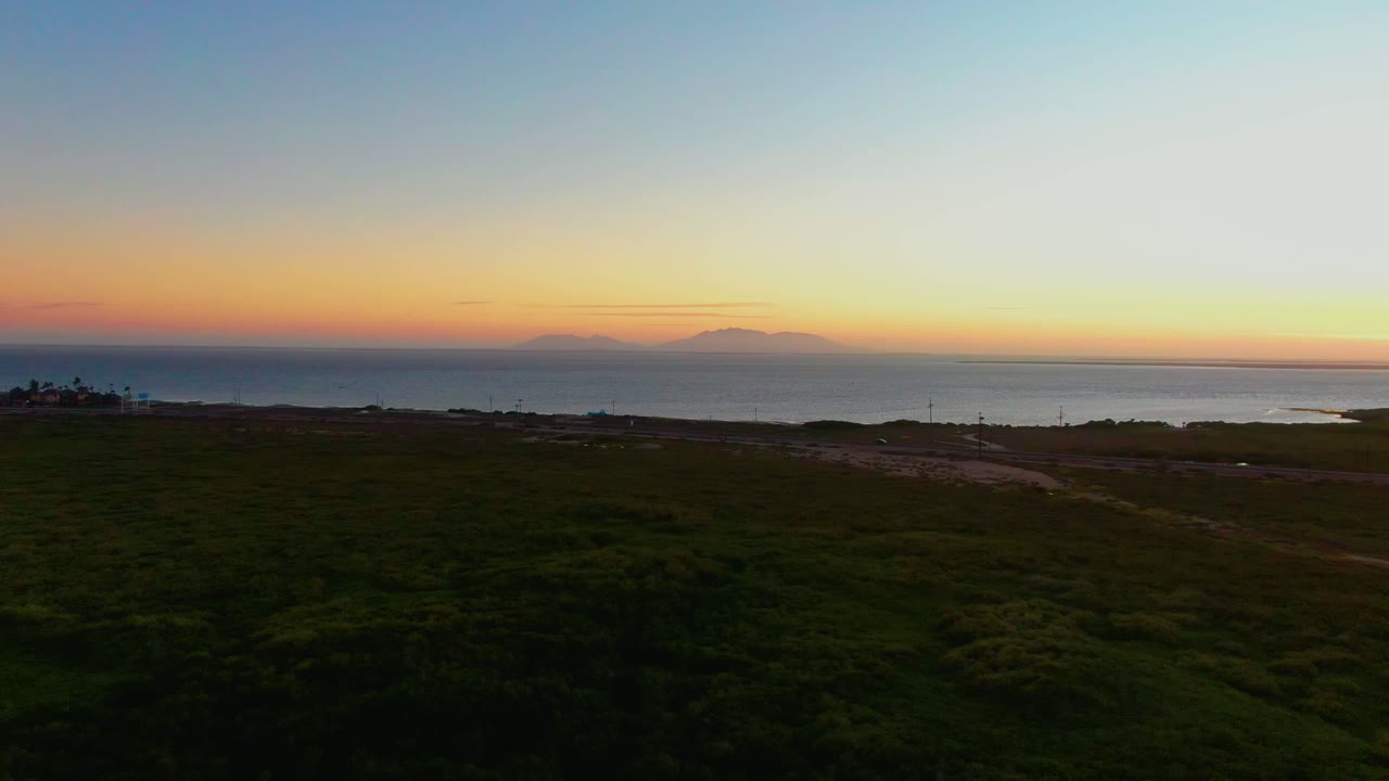Wide aerial-style frame of San Carlos coastal grassland with low shoreline terrain, ocean water, and a narrow orange sunset band beneath pale blue sky, no visible people.