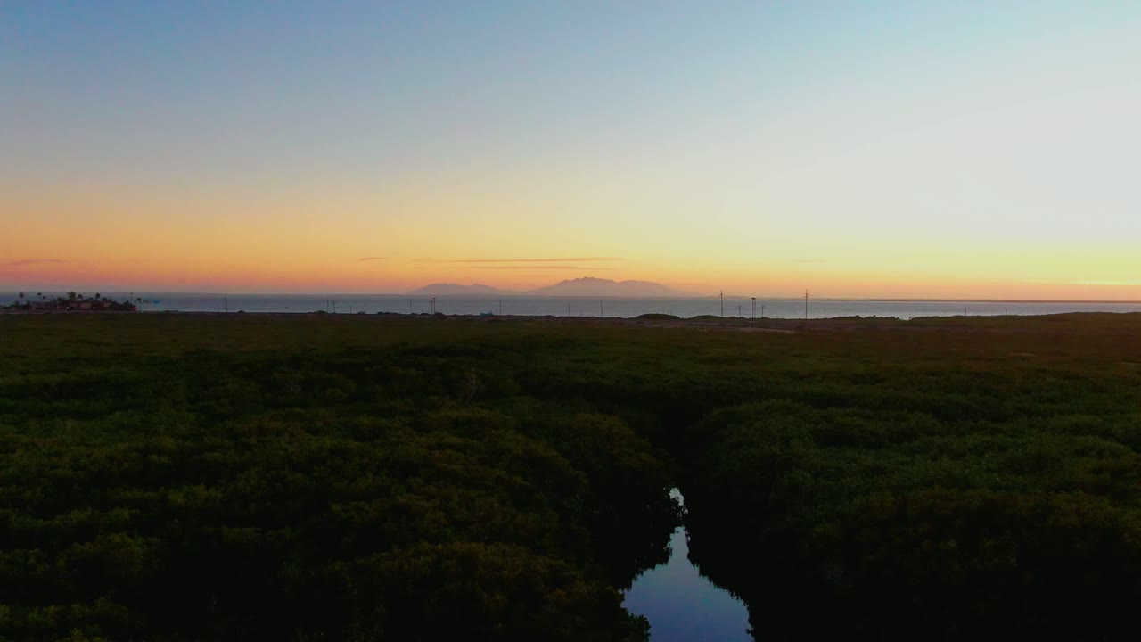 Wide aerial view of dense vegetation, reflective water, and orange sunset horizon.