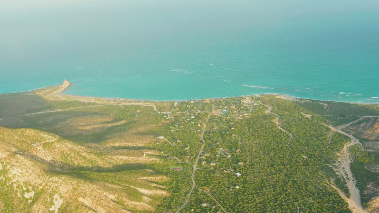 High aerial view over Cabo Pulmo coastal hills toward a pale beach and turquoise ocean, with scattered structures and no visible people.