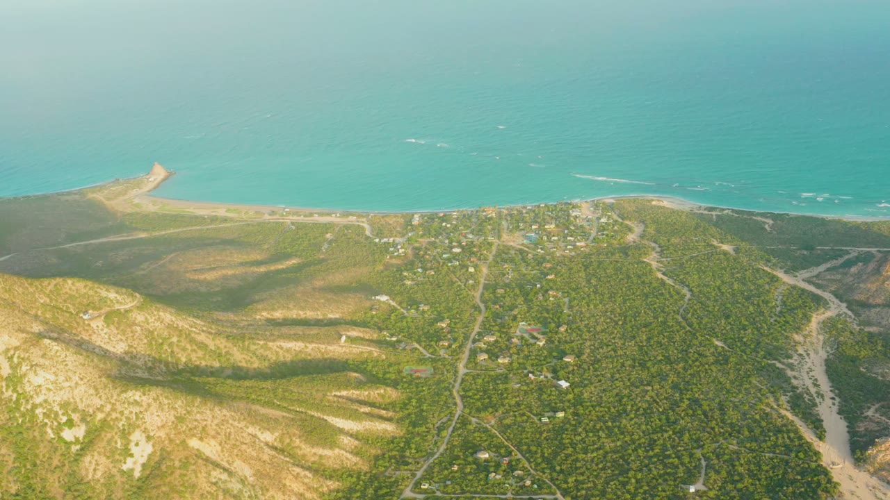 Wide Cabo Pulmo aerial with scrubby hills in the foreground, small structures near the shore, light wave marks offshore, and no visible people.