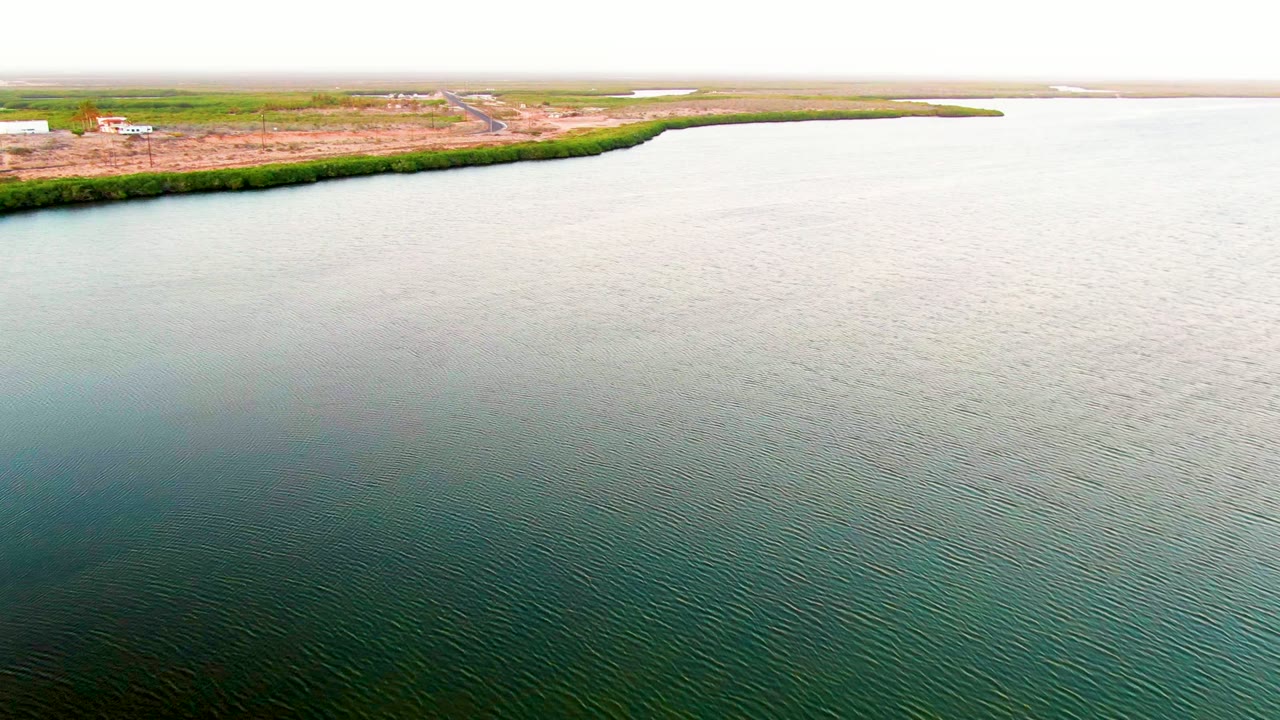 Aerial view in San Carlos, Baja California Sur, with rippling blue-green water below, a green shoreline strip, cleared land, small structures, and no visible people.