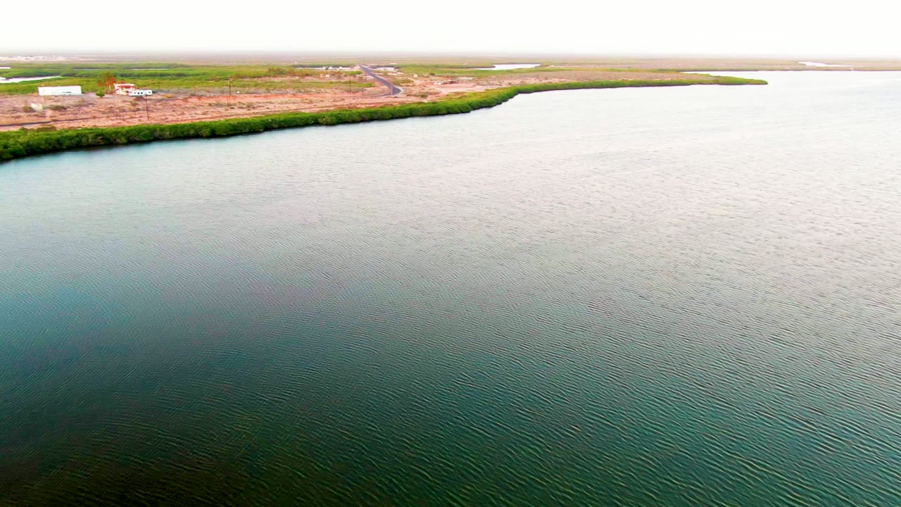 Wide aerial frame in San Carlos with textured water occupying the foreground, a dark green shoreline band, pale cleared ground, scattered structures, and no visible people.